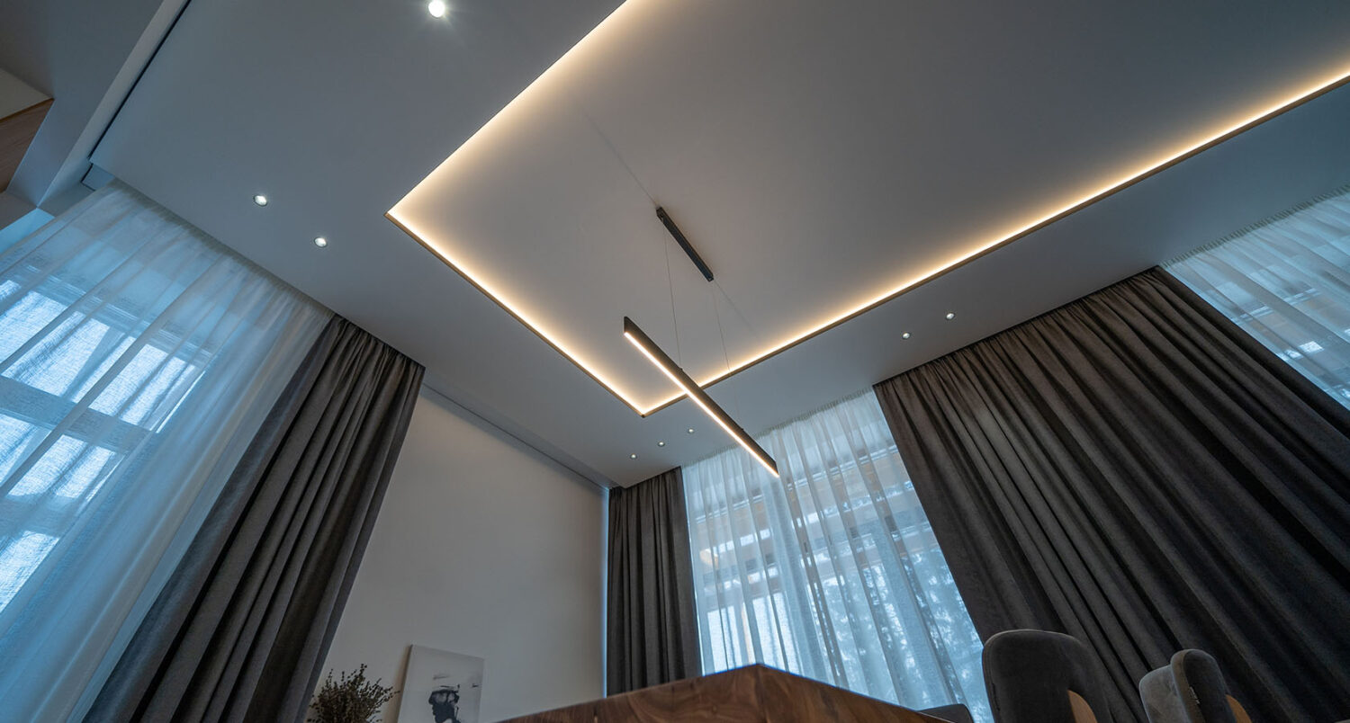 Tavan extensibil alb cu bandă LED perimetrală caldă și corp de iluminat liniar suspendat, într-un living cu draperii gri